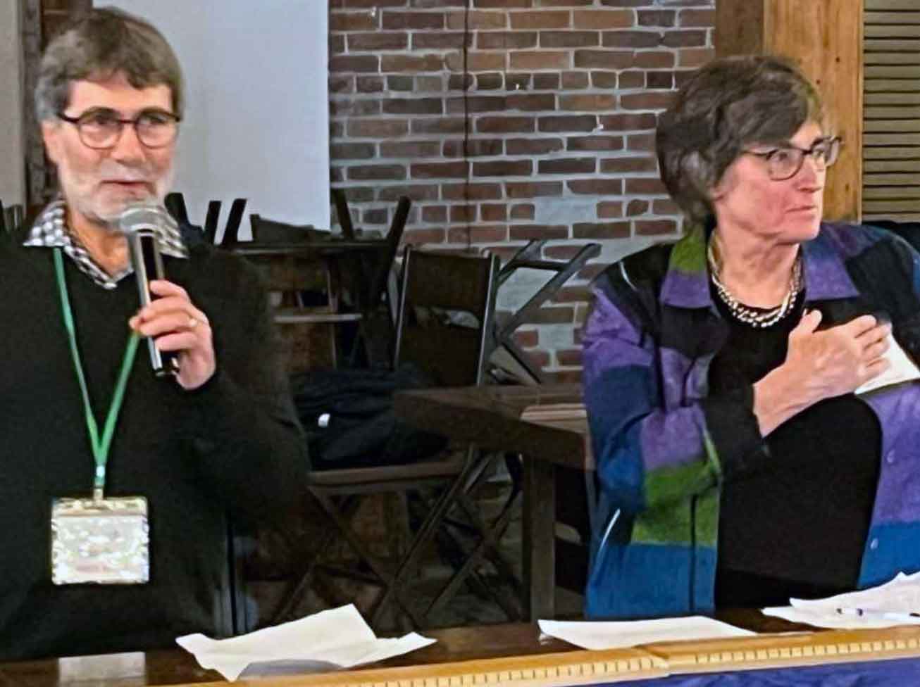 two seated people at a table. One person is holding a microphone.